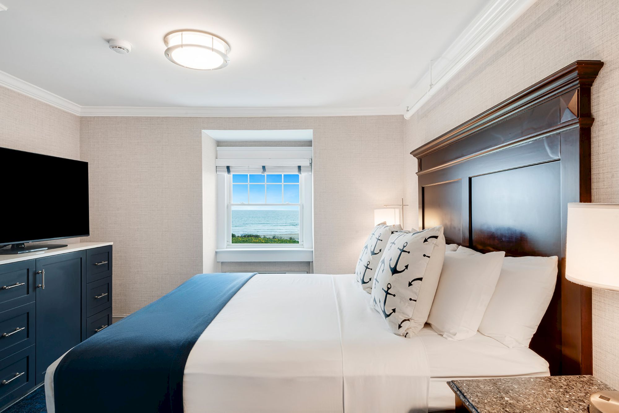 A cozy bedroom with a large bed, decorative pillows, a flat-screen TV, and a window offering an ocean view in the background.