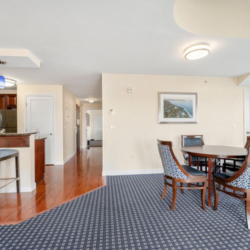 The image shows a modern open-plan dining and kitchen area with a round table, patterned chairs, and a kitchen counter with bar stools.