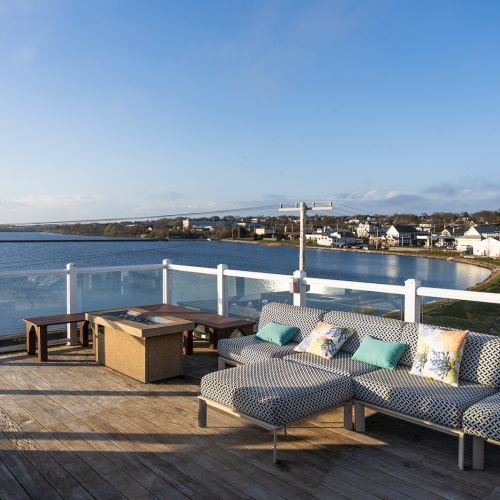 A rooftop patio with seating overlooks a body of water and a small town in the distance under a clear blue sky.