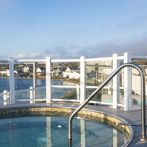 A hot tub with a metal handrail on a deck, surrounded by glass panels with a scenic water and buildings view in the background under a clear sky.