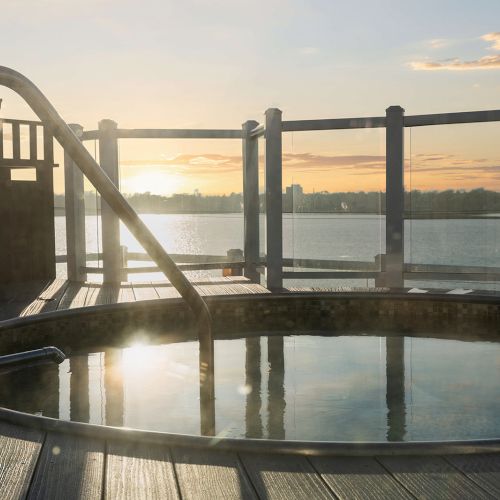 An outdoor hot tub on a wooden deck overlooking a calm lake at sunset.