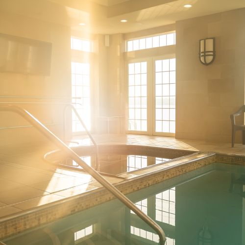 An indoor pool area with large windows, a chair, and a pool. Sunlight is streaming in, creating a warm and inviting atmosphere, ending the sentence.