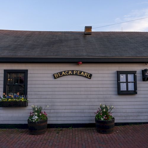 A quaint building with a sign that reads "BLACK PEARL" above the door, along with potted plants and flowers on a brick walkway.