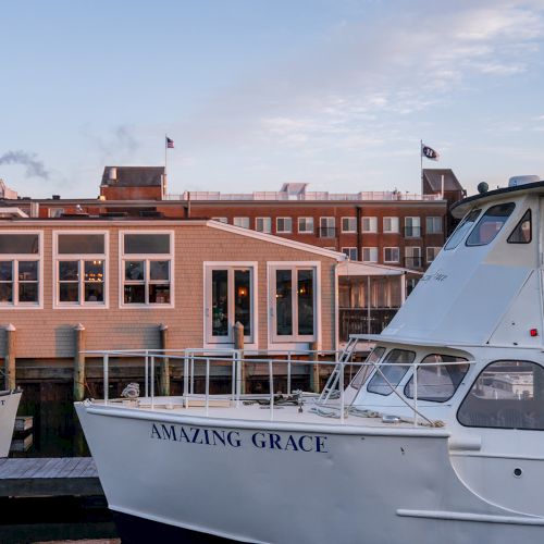 A white boat named "Amazing Grace" is docked by a waterfront with a building in the background under a partly cloudy sky.