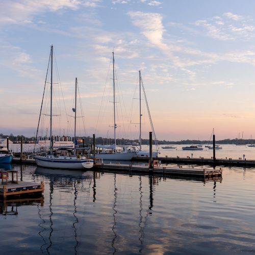 The image shows a serene marina with sailboats docked, calm water reflecting the sky, and an orange-tinted sunset in the background.