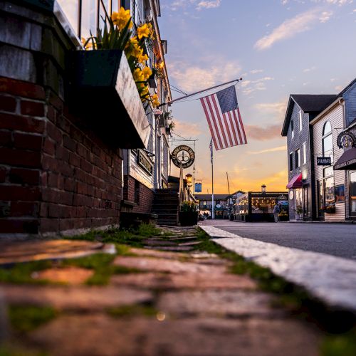 A brick sidewalk leads to a street with buildings, a flag waving, and a sunset in the background, creating a quaint and peaceful scene.