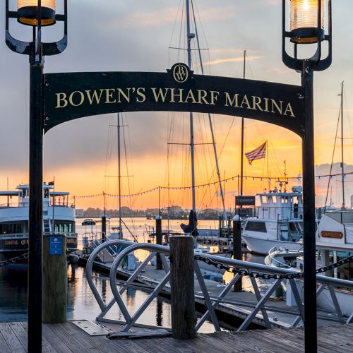 The image features an entrance to Bowen's Wharf Marina with a sign, lamps, and boats in the background, against a sunset sky.