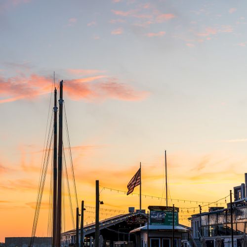 A boat is docked at a pier during sunset, with an American flag flying. String lights add a warm ambiance to the scene.