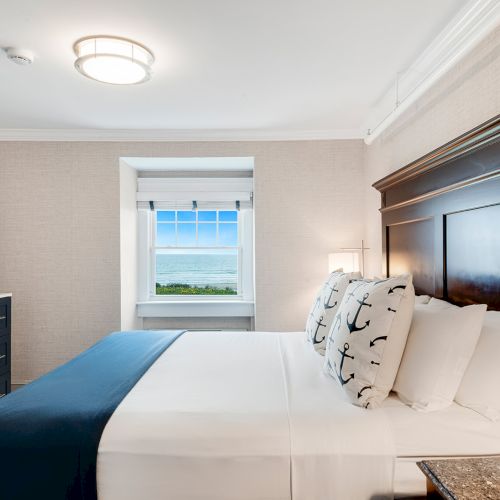 A neatly made bed with nautical-themed pillows is in a room with a TV, dresser, and window offering an ocean view, under soft ceiling lighting.