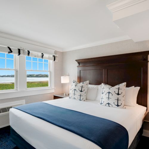 A well-lit bedroom features a large bed with navy and white bedding, anchor-themed pillows, nightstands with lamps, and a window revealing a scenic water view.