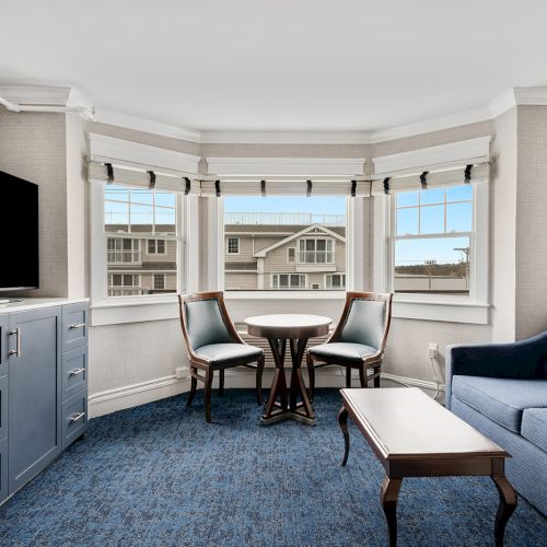 A cozy living room with a blue carpet, a sofa, a small table with two chairs, a TV on a cabinet, and windows showing a view of neighboring buildings.