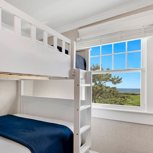 A room with white bunk beds, blue bedding, a large window offering a scenic view of greenery and a sea or ocean in the distance.