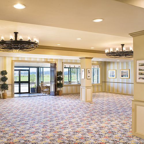 A spacious, brightly lit room with patterned carpet, large windows, and chandeliers. Pillars are decorated with framed pictures, and plants adorn the corners.