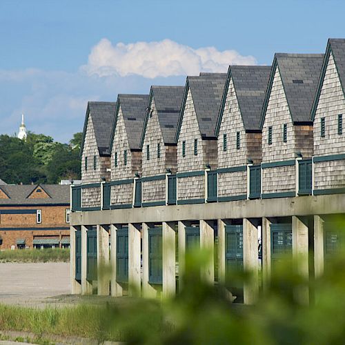 The image shows a row of beachside cottages on stilts, with a sandy beach and buildings, including a church tower, in the background.