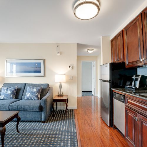 A well-lit living space with a blue couch, coffee table, framed picture, kitchenette with wooden cabinets, and corridor leading to other rooms.