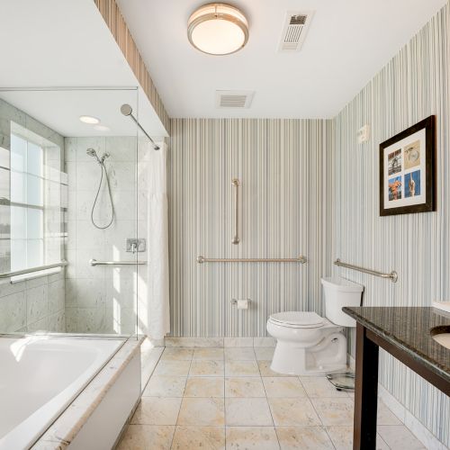 A modern bathroom with a bathtub, walk-in shower, toilet, sink, striped wallpaper, large mirror, grab bars, and a window with a scenic view.