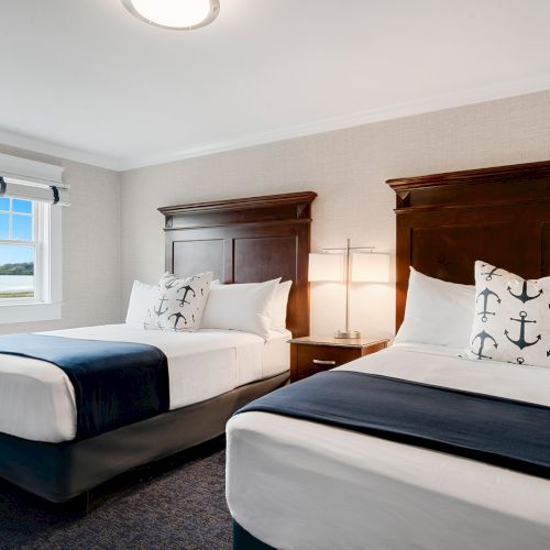 A hotel room features two neatly made beds with white linens and navy blue accents, a window with a view, and nautical-themed pillows.