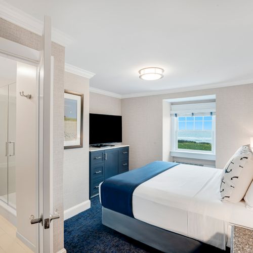 A well-lit hotel room featuring a bed, a TV, and an ocean view through the window. An adjoining bathroom with a shower is also visible.