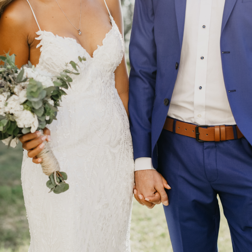 A bride in a white lace dress holding a bouquet and a groom in a blue suit hold hands outdoors, making a wedding moment.