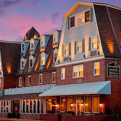 A multi-story brick building with a sign saying "Newport Beach Hotel & Suites" at dusk, warmly lit and featuring multiple dormer windows.