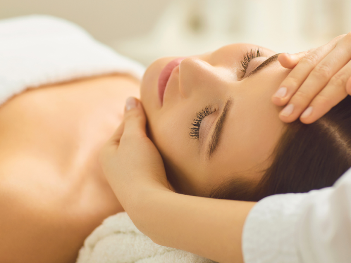 A person is lying down with eyes closed, receiving a facial massage from another person, set in a calm and relaxing environment.