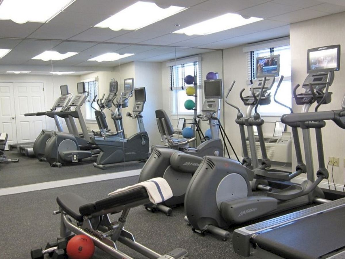 The image shows a gym with various exercise equipment, including treadmills, ellipticals, exercise balls, and weights. There's a mirrored wall and a TV.