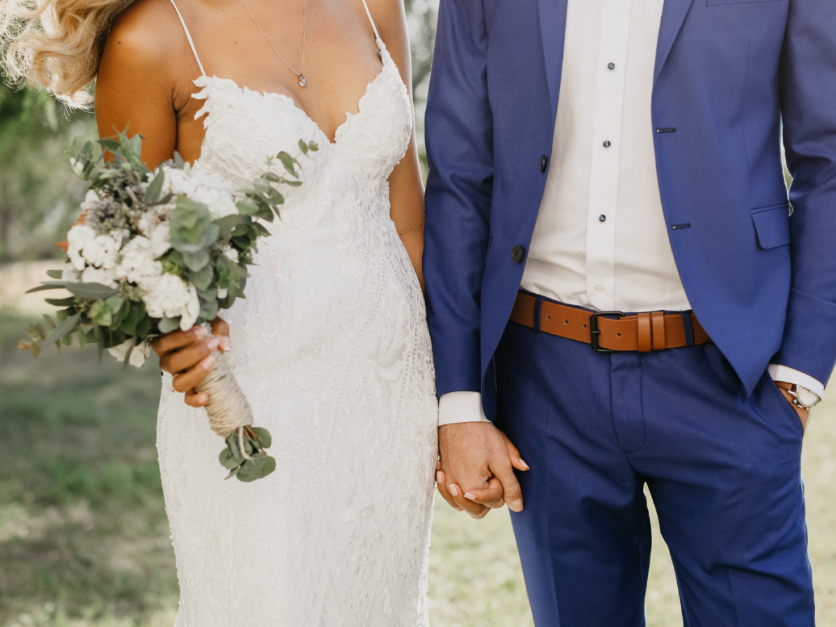 A couple is holding hands outdoors, dressed in wedding attire: the bride in a white dress holding a bouquet, the groom in a blue suit with a brown belt.