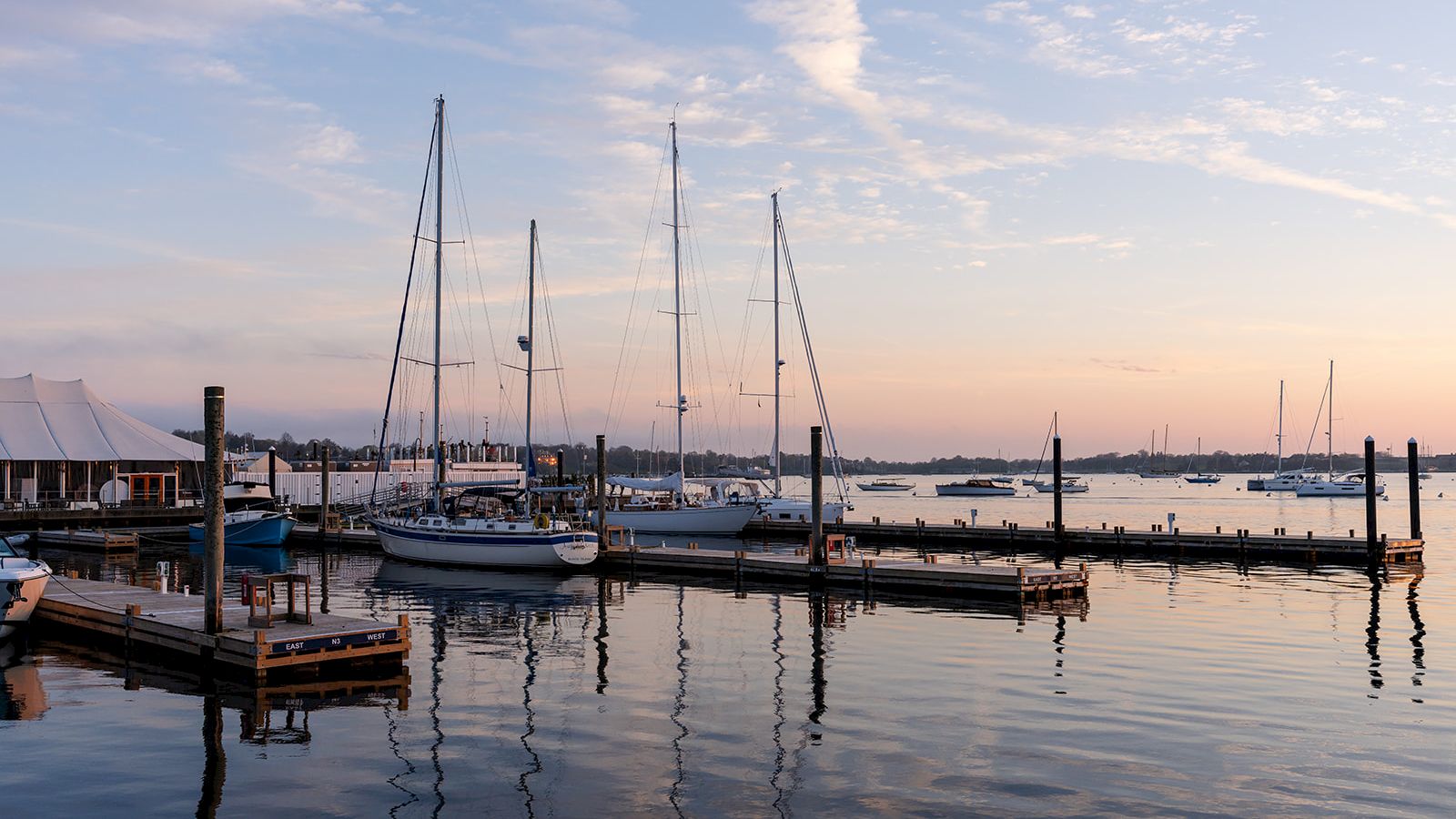 The image depicts a serene harbor scene with sailboats docked at a pier during sunset with a calm water reflection.