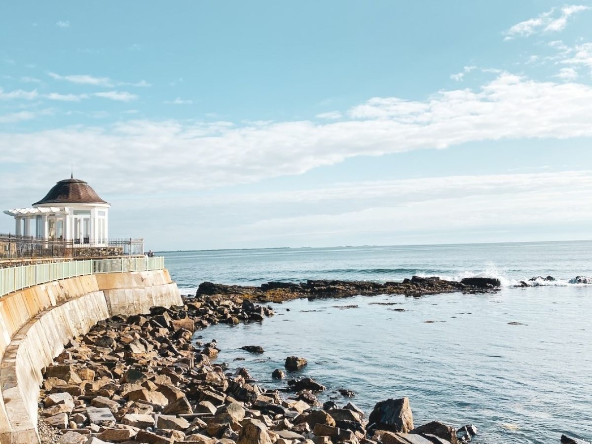 A scenic view featuring a rocky shoreline, a calm sea, and a small gazebo-like structure on the left side under a blue sky with scattered clouds.