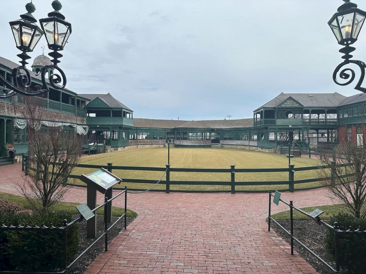 This image shows a courtyard with a grassy area surrounded by fencing, brick pathways, and buildings with green exteriors and lantern-style lights.