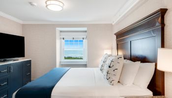 A neatly arranged bedroom with a double bed, nautical-themed pillows, TV, dresser, and a window offering a scenic ocean view ends the sentence.