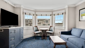 A cozy room featuring a blue couch, a TV on a cabinet, a coffee table, and a small round table with two chairs in front of three large windows.