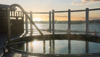 This image shows a circular hot tub on a wooden deck overlooking a body of water as the sun sets, with glass panel railings surrounding them.