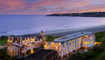 This image shows a coastal area at sunset with illuminated buildings near a beach and the ocean in the background, taken from an elevated viewpoint.