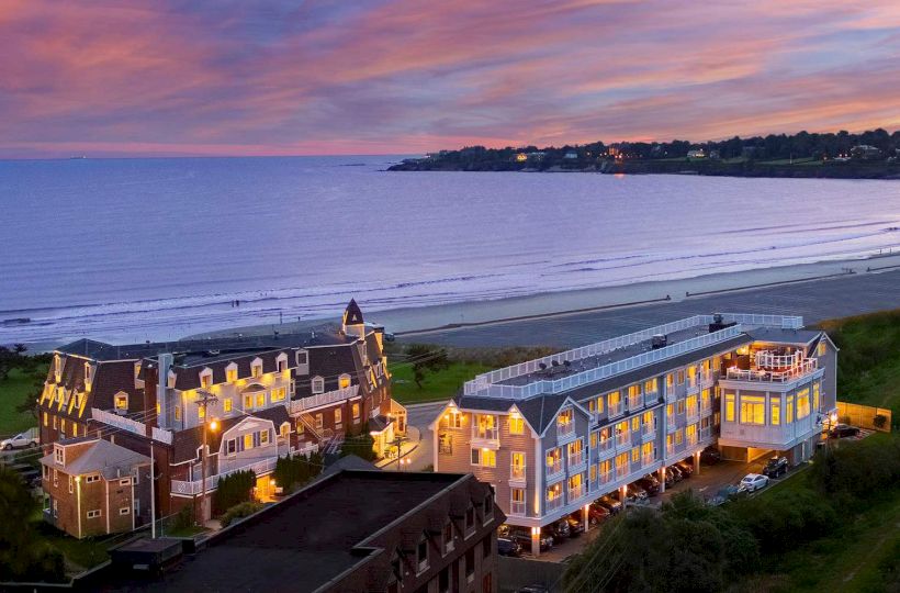 This image shows a coastal area at sunset with illuminated buildings near a beach and the ocean in the background, taken from an elevated viewpoint.