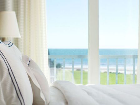 A cozy bedroom with white bedding and a lamp, featuring large windows offering a scenic view of the ocean and a green lawn outside.