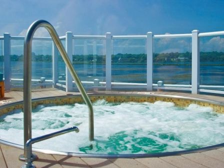 The image shows a round hot tub with bubbling water, positioned on a deck with a clear glass railing, overlooking a body of water in the background.