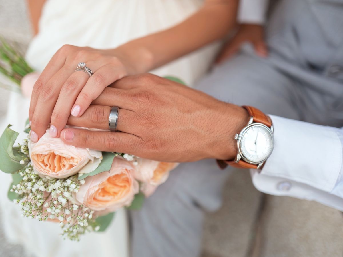 A close-up of a couple's hands, showing wedding rings. They are holding a bouquet of peach roses and baby's breath flowers.