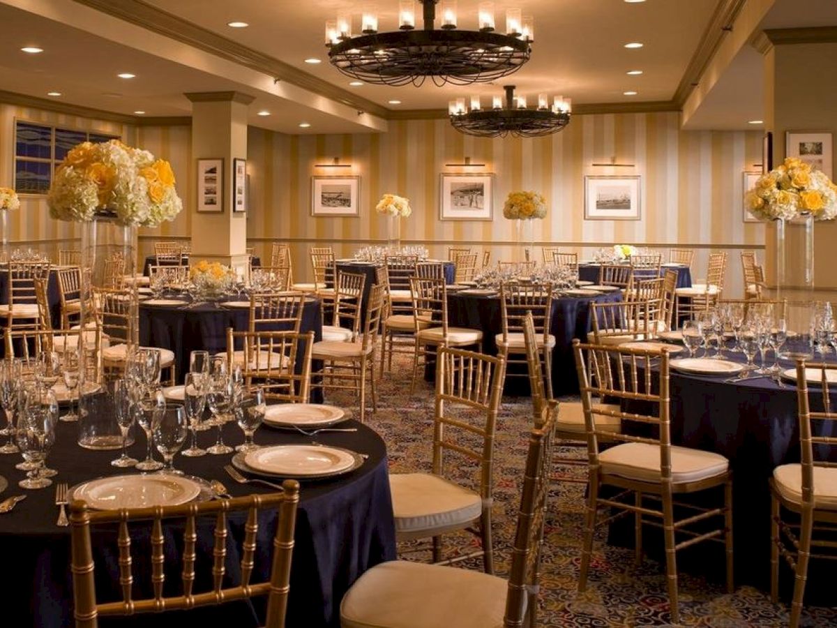 A well-decorated banquet hall with round tables set for an event. Tables have glassware and flower centerpieces, and gold chairs surround them.