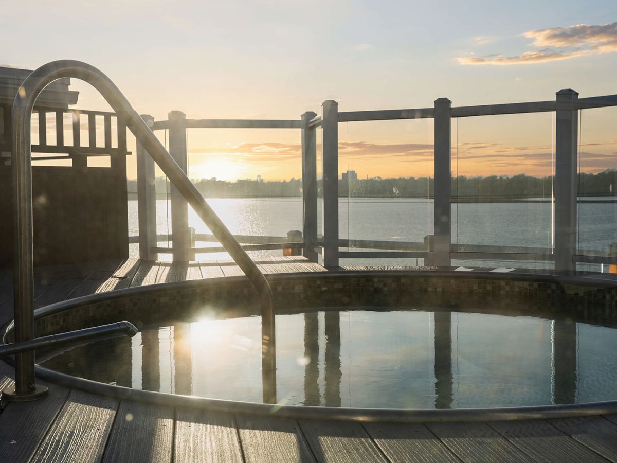 An outdoor hot tub on a wooden deck with a glass railing, overlooking a body of water during sunset.