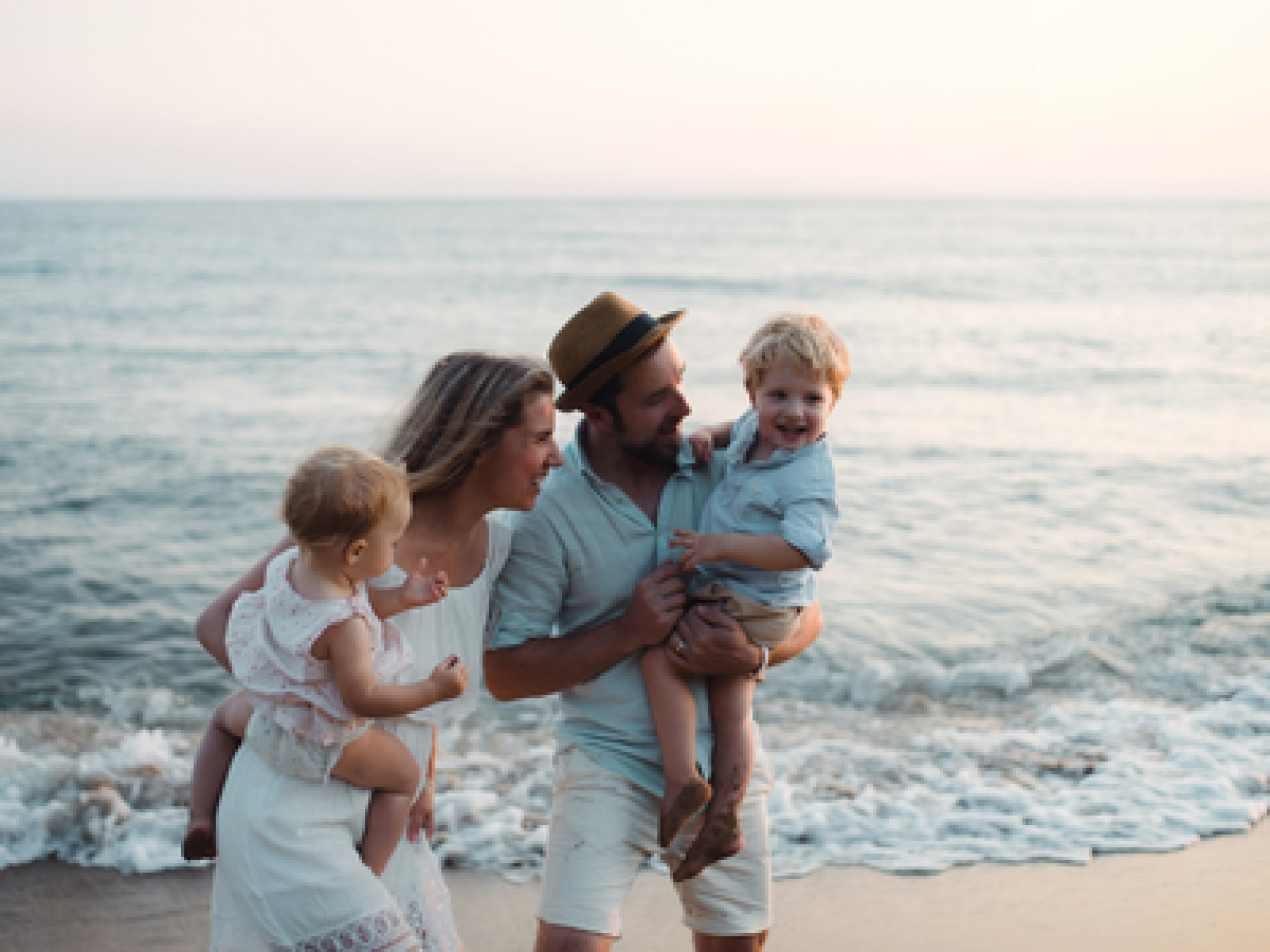 A family with two adults and two children is enjoying a day at the beach, with the parents holding the children by the shoreline as the sun sets.