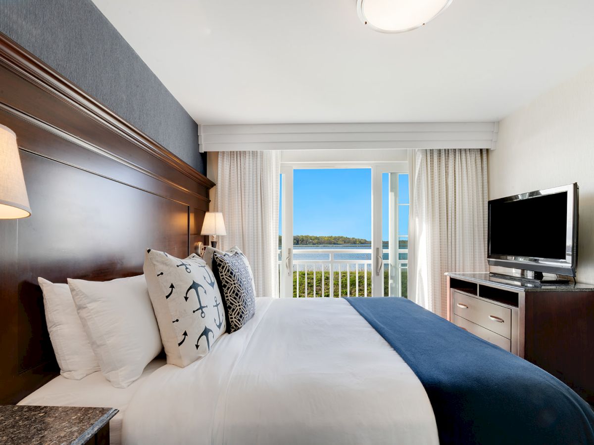 A cozy hotel room with a large bed, pillows, a lamp, a TV on a dresser, and a balcony view overlooking a serene landscape and water.