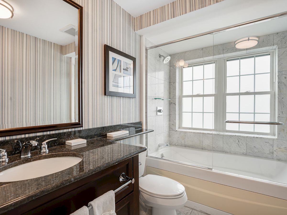 A modern bathroom featuring a sink with a granite countertop, a toilet, and a bathtub with a shower surrounded by marble tiles, and a large mirror.