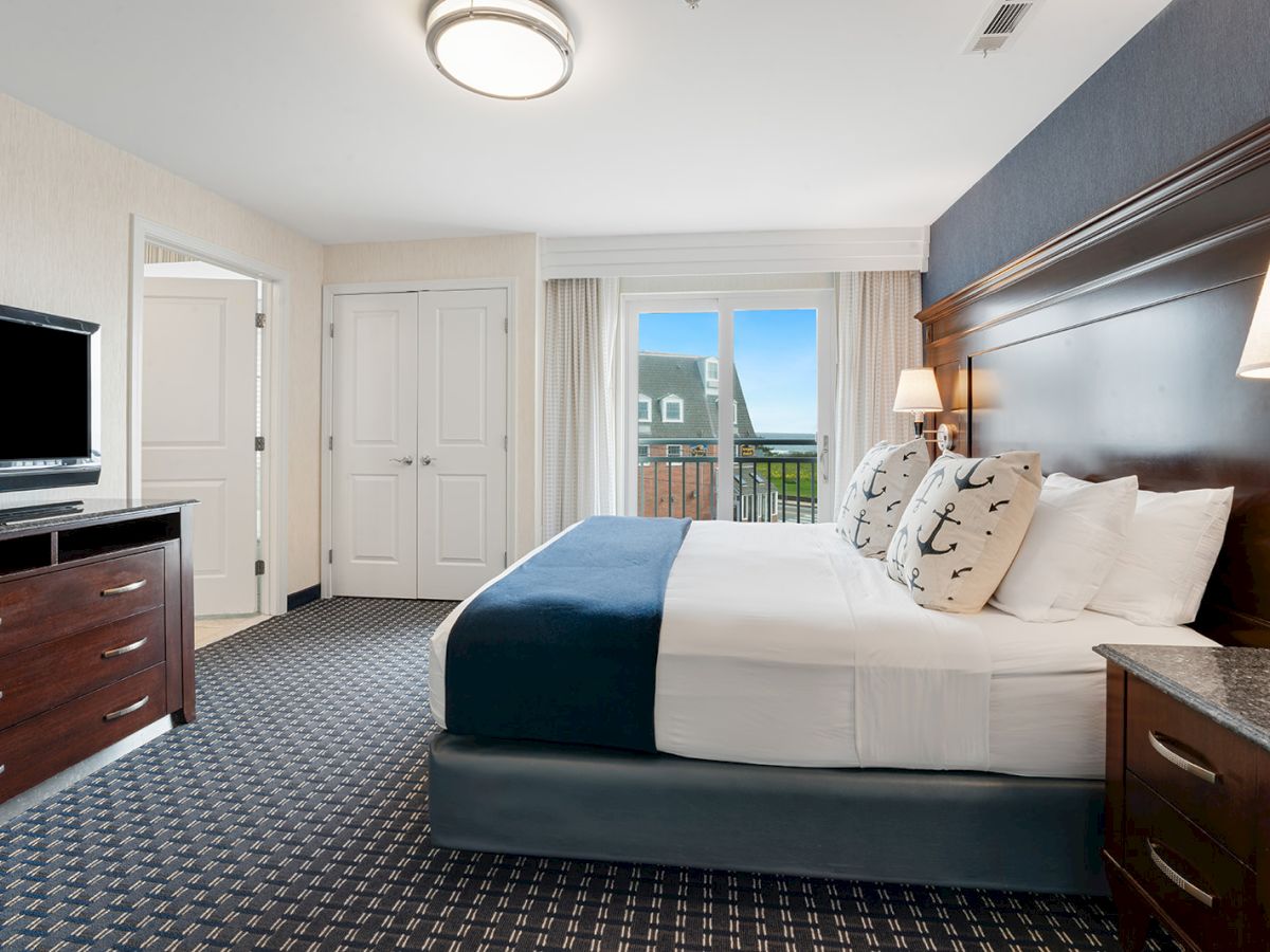 This image shows a hotel room with a king-size bed, a TV on a dresser, and two bedside tables, with a window leading to a balcony.