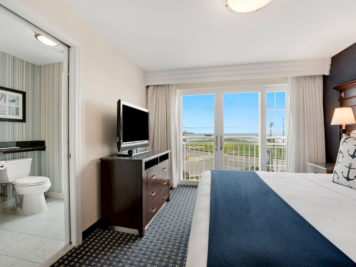 A hotel room with a bathroom, a TV on a dresser, a bed, and a balcony with a view of the ocean. The room is decorated in neutral tones.