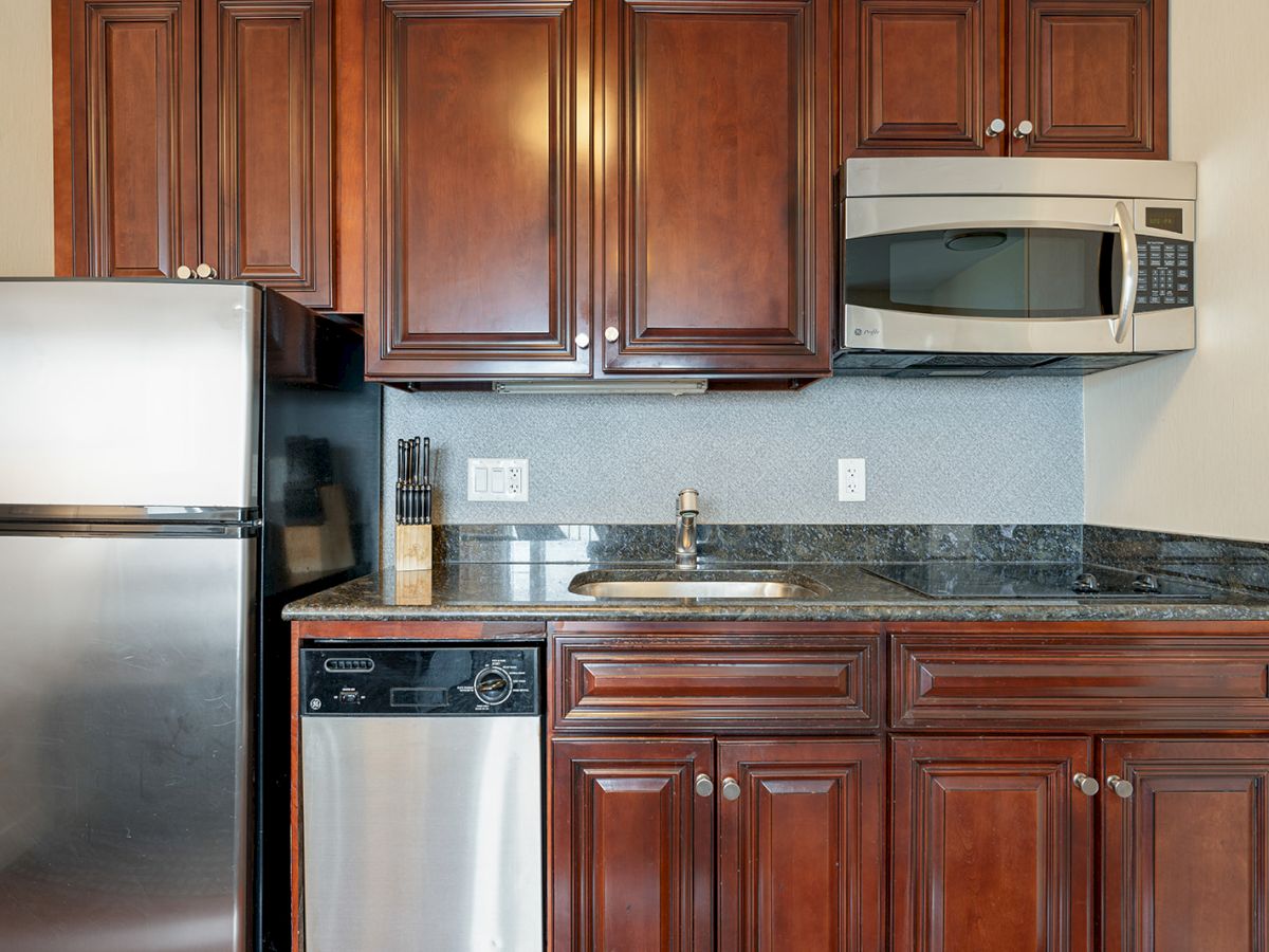 A modern kitchen with wooden cabinets, a stainless steel refrigerator, dishwasher, microwave, and a sink on a granite countertop.