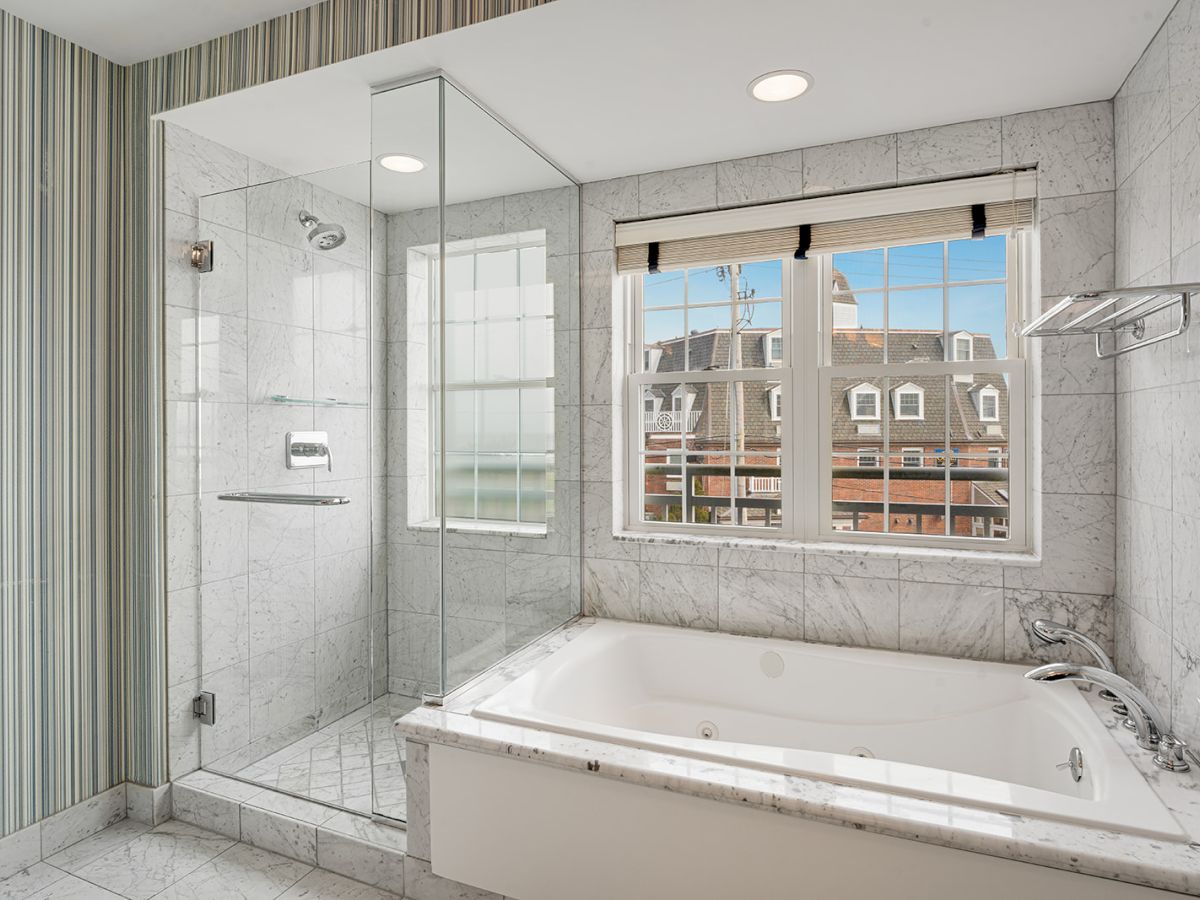 The image shows a modern bathroom with a glass-enclosed shower, a large bathtub, and windows providing a view of buildings outside.