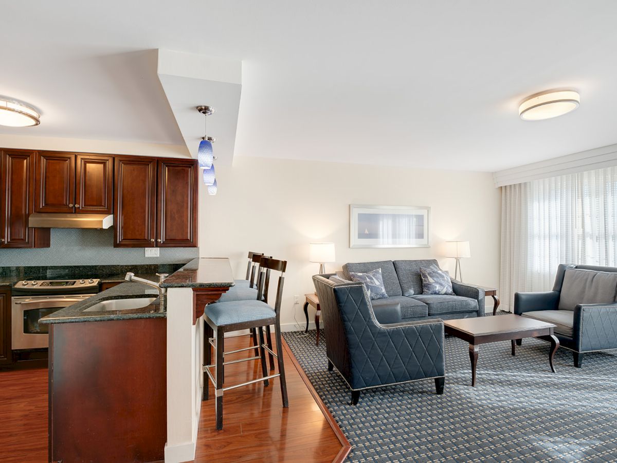 A modern living room and kitchen area with dark wood cabinetry, bar stools, a sectional sofa, armchairs, a coffee table, and a large window with curtains.
