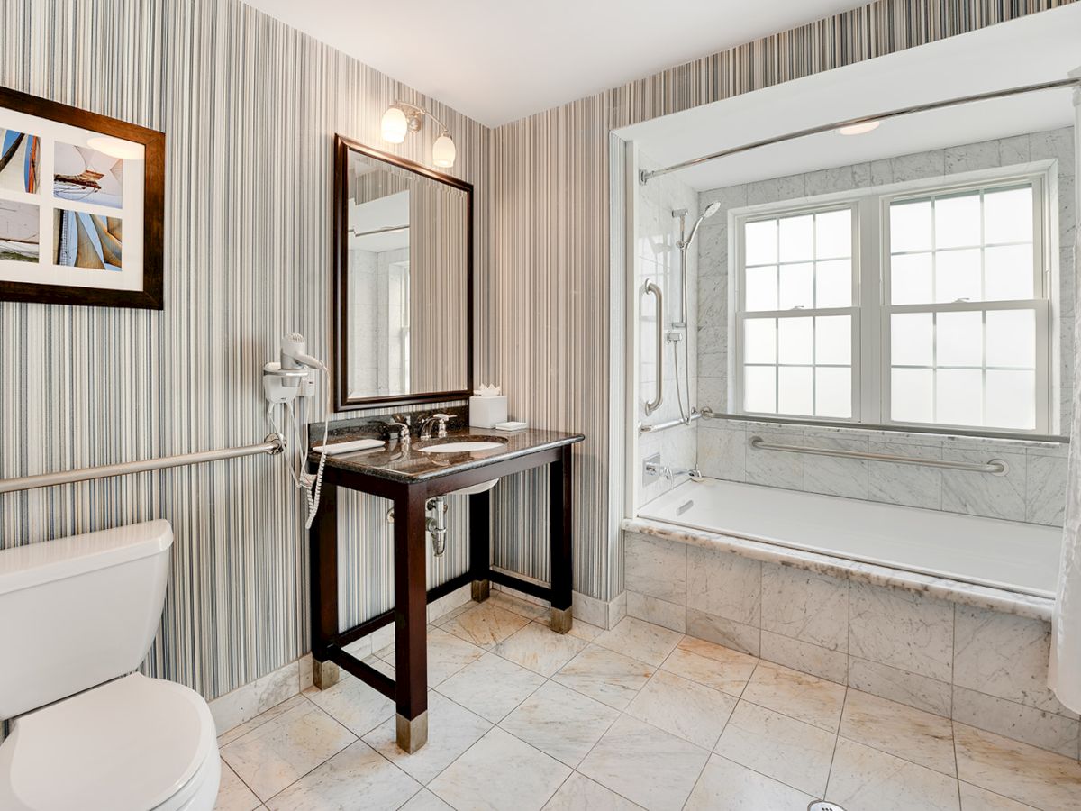 A modern bathroom with a toilet, sink with a mirror, striped wallpaper, framed artwork, and a large bathtub under a window.