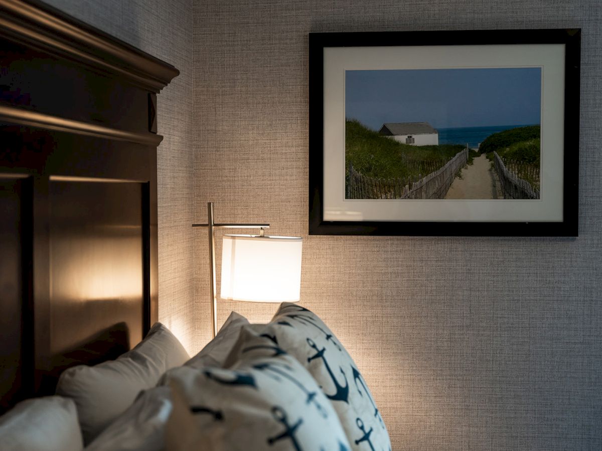 A cozy bedroom corner with a wooden headboard, nautical-themed pillows, a bedside lamp, and artwork depicting a coastal path hanging on the wall.